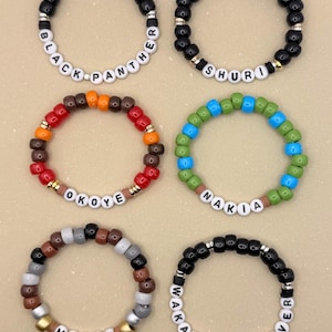 May include: Six beaded bracelets with white letter beads spelling "BLACK PANTHER", "SHURI", "OKOYE", "NAKIA", "MBAKU", and "WAKANDA FOREVER". The bracelets feature black, brown, red, orange, blue, green, gray, and gold beads.