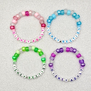 May include: Four beaded bracelets in pink, blue, green, and purple. Each bracelet has clear beads and colorful accent beads. The bracelets spell out "DISNEY ADULT" and "DISNEY BESTIES" with white letter beads.