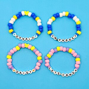 May include: Four colorful beaded bracelets with white letter beads spelling out "DONALD", "AW PHOOEY", "DAISY", and "LETS CONGA". The bracelets feature yellow, blue, pink, and purple beads, with a cartoon character accent bead. Set against a blue background.