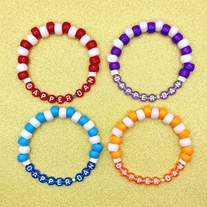 May include: Four beaded bracelets in red, purple, blue, and orange. Each bracelet has white beads and beads that spell out "DAPPER DAN". The bracelets are on a yellow background.