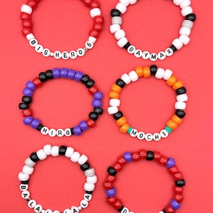 May include: Six colorful beaded bracelets with white letter beads spelling out "BIG HERO 6", "BAYMAX", "HIRO", "MOCHI", "BALALALA", and "BAYMAX". The bracelets are red, white, black, purple, orange, and green.
