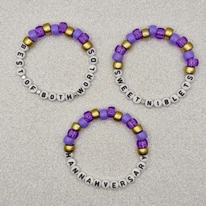 May include: Three beaded bracelets with white letter beads spelling out phrases. The bracelets feature purple, gold, and white beads. One bracelet says "BEST OF BOTH WORLDS", another says "SWEET NIBBLETS", and the third says "HANNAHVERSARY".