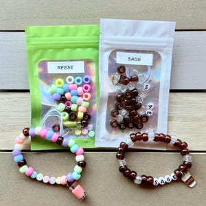 May include: Two clear plastic bags with colorful beads and a charm. The bags are labeled "Reese" and "Sage". Two bracelets are shown, one with brown and white beads and the other with brown, white, and pastel beads. Both bracelets have a charm attached.
