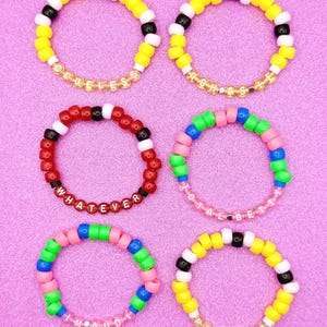 May include: Six colorful beaded bracelets with various phrases. Yellow, black, and white bracelets spell "Clueless" and "High School". A red bracelet says "Whatever". Other bracelets are pink, green, and blue, with the phrases "I'm a dude" and "Total Betty" in gold letters.