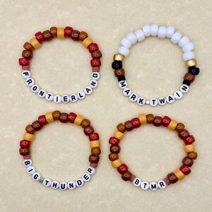 May include: Four beaded bracelets. Two bracelets have the text "FRONTIERLAND" and "BIG THUNDER". The other two bracelets have the text "MARK TWAIN" and "BTMR". The bracelets are made of colorful beads in shades of red, brown, yellow, and white.