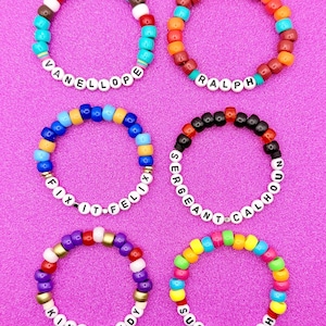 May include: Six colorful beaded bracelets with names spelled out in white letter beads. The bracelets are in various colors, including turquoise, orange, blue, purple, and yellow. Names include Vanellope, Ralph, Fix-It-Felix, Sergeant Calhoun, King Candy, and Sugar Rush.