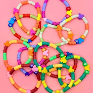 May include: A collection of colorful beaded bracelets in various shades of red, orange, yellow, green, blue, and purple. Each bracelet features a unique design with decorative accents, including small, shaped beads. The bracelets are arranged on a pink background.