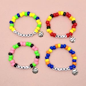 May include: Four colorful beaded bracelets with white letter beads spelling out names: JACKSON, WOLF, SKOLE, and WATSON. Each bracelet has a baseball charm. The beads are in various colors, including yellow, blue, green, pink, orange, and red.