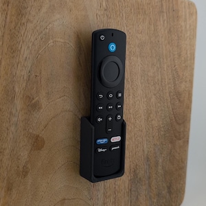 以下が含まれることがあります： 円形のボタンと、ナビゲーション、音量、ストリーミングサービス（Netflix、Disney+、Prime Video、Peacockなど）用のボタンがいくつかある黒いリモコン。リモコンは黒いホルダーで壁に取り付けられています。