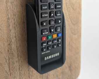 May include: A black Samsung TV remote control with a variety of buttons, including power, volume, channel, and input selection. The remote is mounted on a wooden wall mount.