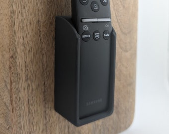 Może przedstawiać: Czarny pilot do telewizora Samsung z uchwytem przymocowanym do drewnianej powierzchni. Pilot ma przyciski do Netflix, Prime Video i Hulu.