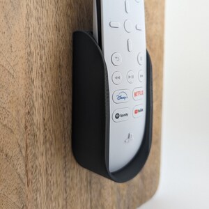 May include: A black remote control holder mounted on a wooden surface. The holder is designed to hold a white remote control with buttons for Netflix, Disney+, Spotify, and YouTube.