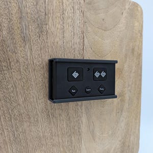 May include: A rectangular black electronic control device with five buttons mounted on a light brown wood surface. The top row features two square buttons with geometric logos, while the bottom row has three diamond-shaped buttons labeled Test, Learn, and Party.