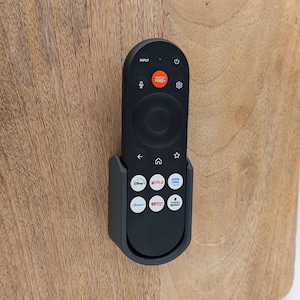 Op de afbeelding: Een zwarte afstandsbediening voor de televisie zit in een donkergrijze wandhouder. De afstandsbediening heeft een cirkelvormig navigatiepaneel, een aan-uitknop en speciale knoppen voor Disney+, Netflix, Prime Video, Paramount+, iHeartRadio en Stingray Music.
