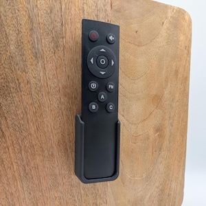 May include: A black remote control with multiple buttons, including power, volume, and function keys, is mounted on a wooden surface. The remote is held in a black holder. The wood has a natural grain pattern.