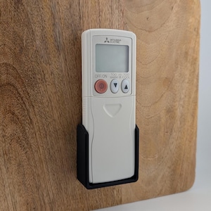 Könnte beinhalten: Eine weiße Mitsubishi Electric Klimaanlagen-Fernbedienung in einer schwarzen Halterung, montiert auf einer Holzoberfläche. Die Fernbedienung hat einen LCD-Bildschirm und Tasten wie "OFF/ON", "TOO WARM" und "TOO COOL".