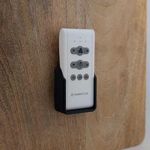 May include: A white ceiling fan remote control with gray buttons and the "FANIMATION" logo, mounted in a black holder on a wooden surface. The remote features buttons for fan speed and light control.
