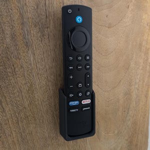以下が含まれることがあります： 円形のナビゲーションパッドと、電源、音量、チャンネルアップとダウン、戻る、ホーム、再生、一時停止、早送り、巻き戻し、マイクのボタンが付いた黒いリモコン。リモコンは黒いホルダーで壁に取り付けられています。ホルダーには、Netflix、Amazon Prime Video、Disney+、Peacockのボタンがあります。