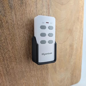 May include: A white remote control with gray buttons and the text "Slyantek" is mounted in a black holder on a wooden surface. The remote has "ON" and "OFF" labels and three numbered buttons.