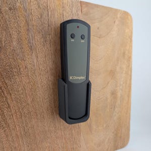 Puede incluir: Un mando a distancia Dimplex negro con soporte, montado sobre una superficie de madera. El mando tiene botones "ON" y "OFF", y una luz indicadora roja. El logotipo de Dimplex es visible.