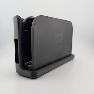 May include: A black Nintendo Switch dock with the words "Nintendo Switch" printed on the front. The dock has a rectangular shape with a slot for the console and a base for stability. The design is sleek and modern.