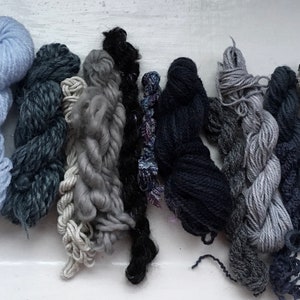May include: An assortment of yarn skeins in various shades of blue, gray, and black. The yarns have different textures and thicknesses, ranging from fine to chunky. The color palette includes light blue, charcoal, and silver.