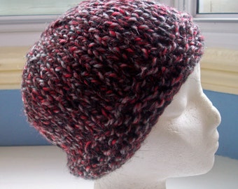 May include: A hand-knitted beanie hat featuring a chunky, variegated yarn pattern in shades of black, dark grey, and vibrant red. The knit texture is thick and consistent throughout the entire surface of the cap, creating a dense, warm appearance.
