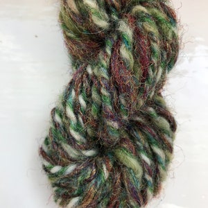 May include: A skein of yarn in a mix of green, brown, white, and blue colors. The yarn is soft and fluffy, and it has a rustic look.