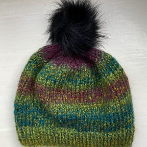 May include: A knitted beanie hat with a black faux fur pom-pom. The hat features a color gradient of green, blue, purple, and yellow. The hat is made of a thick, textured yarn.