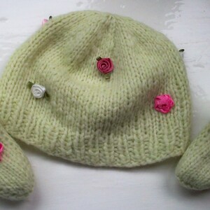 May include: A set of two knitted baby booties and a matching hat. The hat is cream colored with pink and white flower embellishments. The booties are cream colored with pink flower embellishments.