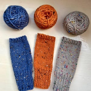 Könnte beinhalten: Drei Knäuel Wolle in Blau, Orange und Grau mit passenden gestrickten Handgelenkwärmern. Die Wolle und die Handgelenkwärmer haben ein gesprenkeltes Design. Die Handgelenkwärmer sind in Blau, Orange und Grau. Die Wolle ist über den Handgelenkwärmern angeordnet.