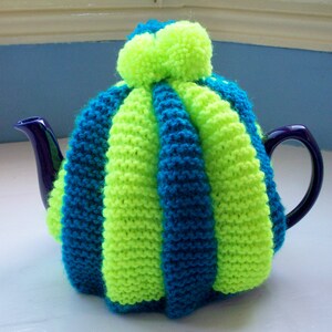 May include: A knitted tea cozy in a vibrant color scheme of blue and neon green stripes. The cozy has a rounded shape with two pom-poms on top. The cozy is designed to fit over a dark blue teapot.