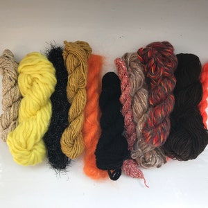 May include: Assortment of yarn skeins in various colors, including yellow, orange, black, brown, and beige. The yarn is arranged in a row on a white surface.