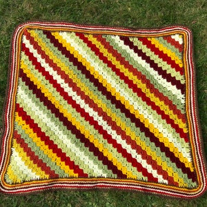May include: A handmade crochet blanket with a diagonal striped pattern in shades of red, yellow, green, and brown. The blanket has a brown border with white and red accents. The blanket is displayed on a green grassy surface.