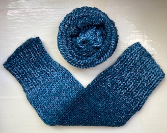 May include: A knitted blue scarf and hat set. The scarf is a long, rectangular shape with a ribbed design, and the hat is a rolled circle. The yarn features a blend of blue hues, giving a textured look. The set is presented on a white surface.