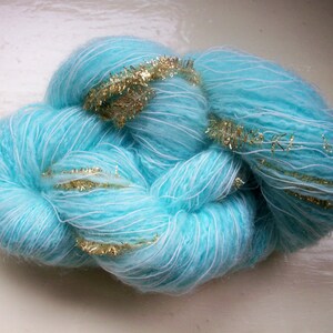 May include: A close-up of a skein of turquoise yarn with white strands and gold tinsel accents. The yarn is fluffy and appears soft, ideal for knitting or crocheting projects. The gold tinsel adds a touch of sparkle.