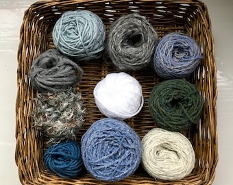 May include: A woven basket filled with various balls of yarn in shades of blue, gray, white, and green. The yarn balls vary in texture and size, creating a visually appealing arrangement. The basket is made of brown woven material.
