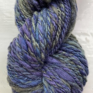 May include: A skein of thick, textured yarn in shades of blue, purple, and gray. The yarn appears to be hand-dyed, with color variations throughout the strands. The yarn is tightly wound, showing the texture of the fibers.