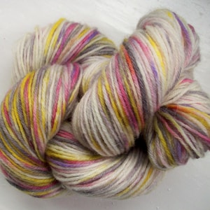 May include: A skein of colorful yarn with a mix of cream, yellow, pink, purple, and gray strands. The yarn is tightly wound, showing the texture and color variations. Ideal for knitting or crocheting projects.