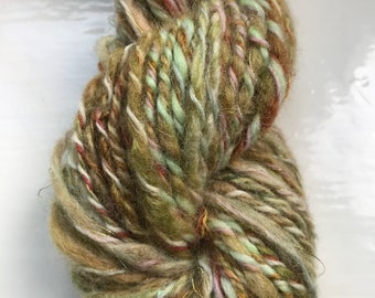 May include: A close-up of a skein of yarn with a mix of colors. The yarn is a blend of greens, browns, and cream, with accents of red, pink, and gold metallic threads. The yarn appears soft and textured, suitable for knitting or crochet projects.