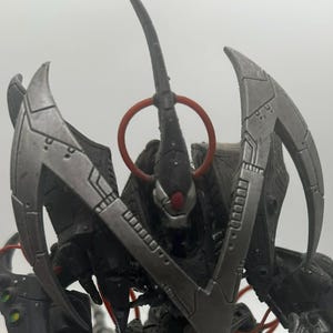May include: A detailed action figure of a dark gray robotic creature with sharp, metallic blades and red accents. The figure has a menacing design, with a large head and multiple articulated limbs, standing on a white surface.