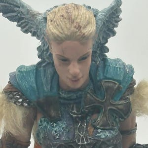 May include: A detailed figurine of a female warrior with blonde hair, wearing a teal and brown outfit with armor and a cross emblem. She holds a staff with intricate detailing, and has feathered wings on her back.