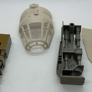 May include: A collection of plastic toy parts including two grey landing gear struts, one olive green landing gear strut, a beige cockpit canopy with a grid window frame, a grey mechanical interior component, and a flat beige panel with molded lines.