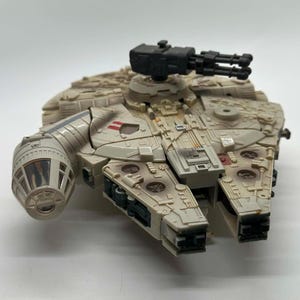 May include: A tan and gray toy spaceship with a black gun turret on top. The spaceship has detailed panels, red accents, and a circular cockpit. The toy is a replica of the Millennium Falcon from the Star Wars franchise.