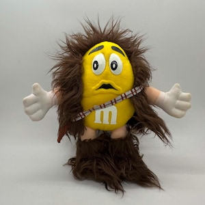 May include: A plush yellow M&M's character dressed as Chewbacca from Star Wars. The toy has brown fur, white hands, and a Chewbacca bandolier. The M&M's logo is visible on the character's belly. The toy has a surprised expression.