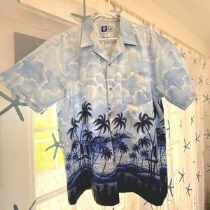 May include: A short-sleeve button-down shirt featuring a light blue sky with white clouds on the upper portion and a dark blue tropical landscape with palm trees and ocean waves on the lower portion. The shirt has a collar and a chest pocket.