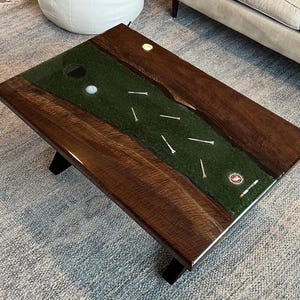 May include: A rectangular coffee table with a dark wood frame and black legs. The center features a green surface with golf tees, a golf ball, and a golf hole. A golf-themed decorative item.