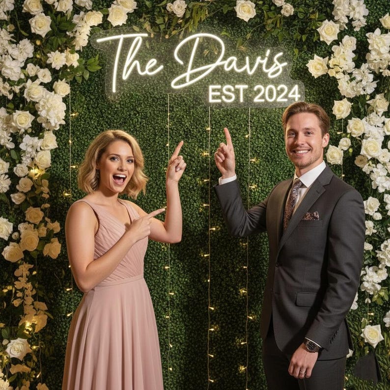 May include: A couple points towards a neon sign reading "The Davis EST 2024". The sign is set against a backdrop of green foliage and white flowers. The woman wears a pink dress, and the man wears a grey suit.