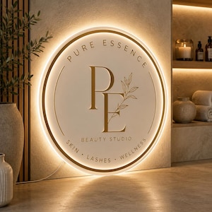 K&ouml;nnte beinhalten: Ein kreisf&ouml;rmiges Wand-Schild mit wei&szlig;er Oberfl&auml;che und goldener Schrift. Der Text lautet PURE ESSENCE, BEAUTY STUDIO sowie SKIN, LASHES, WELLNESS. Ein gro&szlig;es goldenes PE-Monogramm mit Blatt-Detail ist zentriert. Das Schild hat eine warme Hintergrundbeleuchtung.