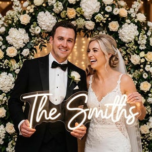 May include: A newly married couple smiling, holding a neon sign that reads "The Smiths." The bride wears a white lace wedding dress, and the groom is in a black tuxedo. They are in front of a floral archway with white and cream flowers.